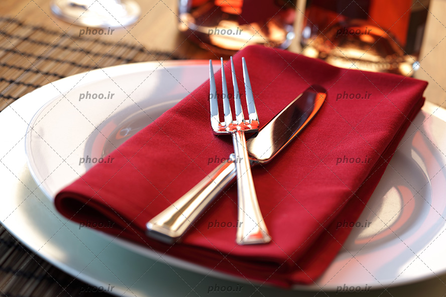 عکس با کیفیت کارد و چنگال بر روی پارچه ای قرمز داخل ظرف چینی سفید بر روی میز رستوران