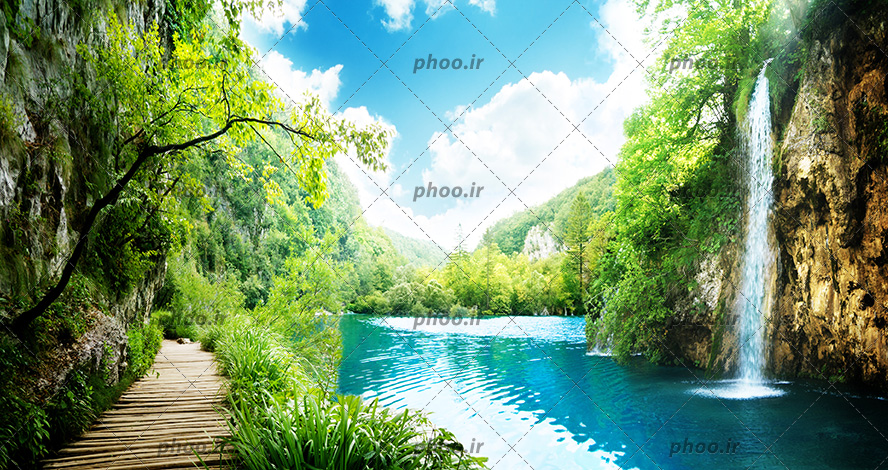عکس با کیفیت پل چوبی در کنار رودخانه زیبا با آب شفاف و آبشار جازی شده از کوه و درختان سرسبز در اطراف پل