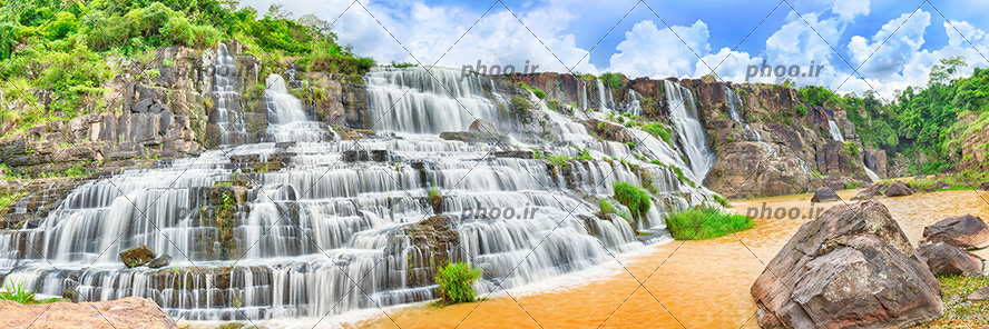 عکس با کیفیت آبشار جاری از بالای کوه های مرتفع و منتهی به دریاچه و آب به رنگ نارنجی و تخته سنگ های بزرگ در کنار دریاچه