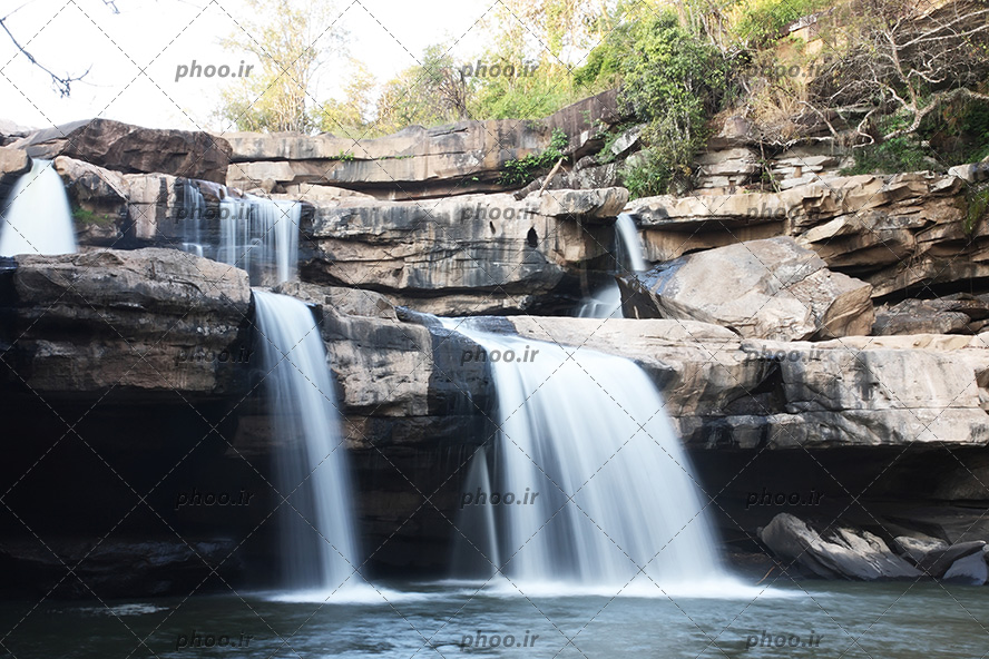 عکس با کیفیت آبشار جاری شده از لا به لای سنگ های بزرگ و شاخه درختان در اطراف صخره ها