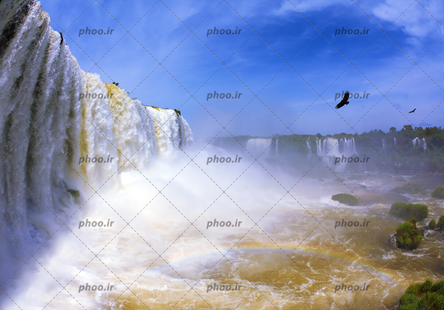 عکس با کیفیت آبشار خروشان و سنگ های بزرگ در دریاچه و پرنده در حال پرواز در آسمان