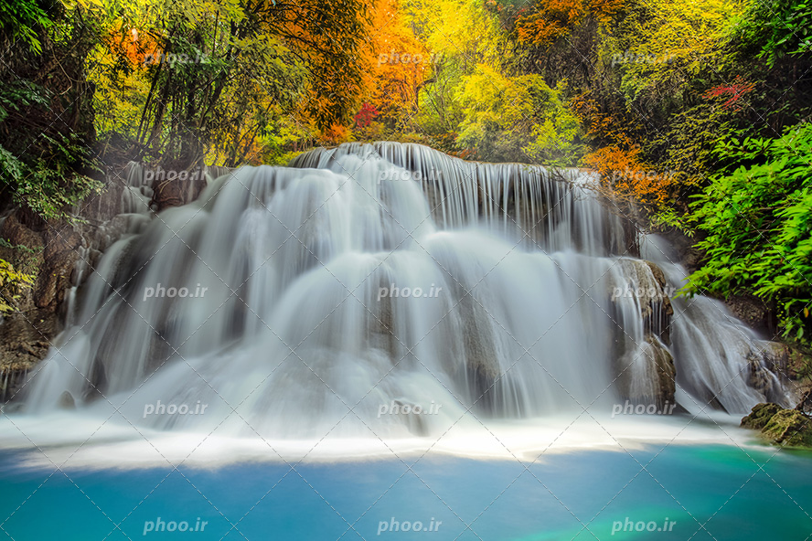 عکس با کیفیت آبشار با آب زلال جاری شده از صخره های بزرگ و درختان پاییزی در اطراف رودخانه