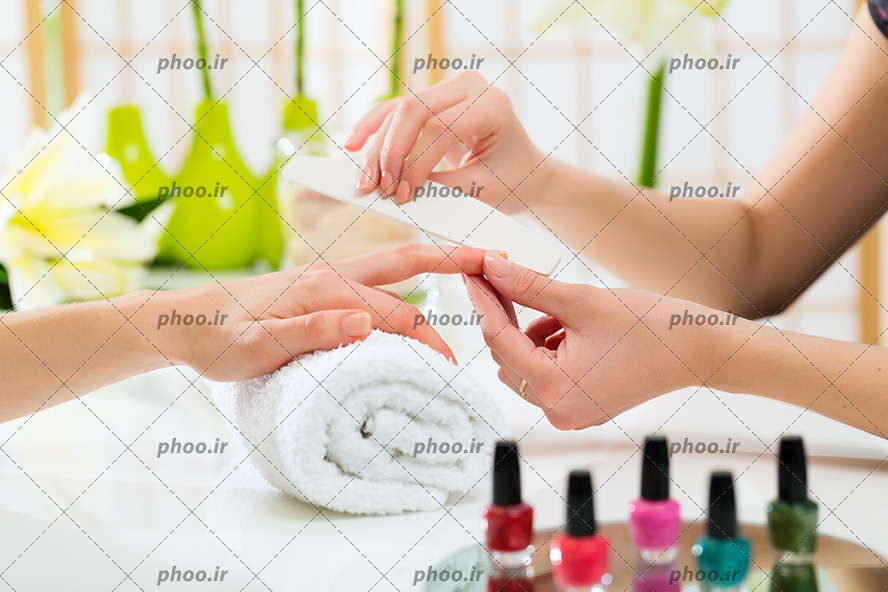 عکس با کیفیت آرایشگر در حال سوهان کردن ناخن های زن و لاک های رنگارنگ در کنار یکدیگر چیده شده بر روی میز