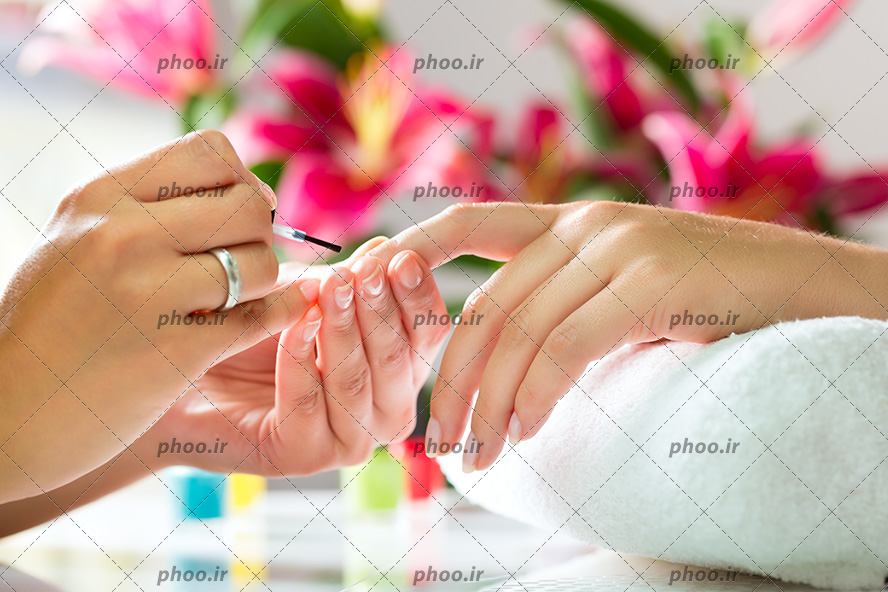 عکس با کیفیت زن آرایشگر در حال لاک زدن ناخن های مشتری و در کنار دست آنها گل های زیبا و لاک های رنگارنگ چیده شده بر روی میز