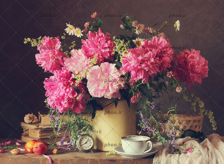 عکس با کیفیت فنجان و ساعت کوچک و چند کتاب و دو سیب در کنار گلدان پر شده از گل های زیبا به رنگ صورتی