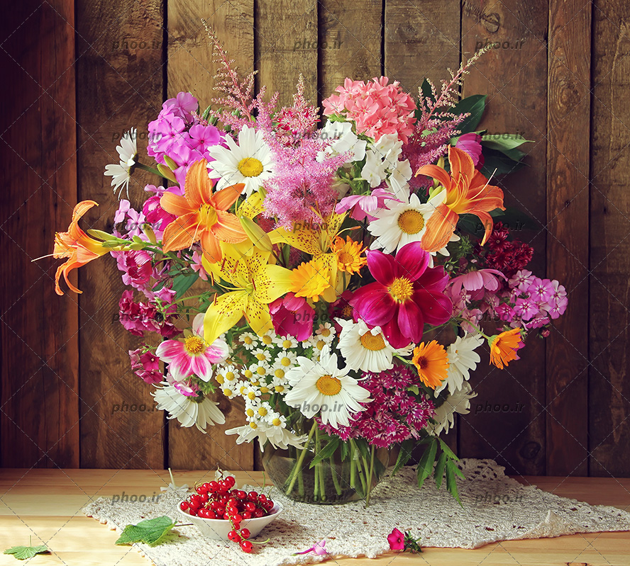 عکس با کیفیت انواع گل رنگارنگ در گلدان بلوری روی میز چوبی و گیلاس های تازه داخل ظرف و دیوار چوبی