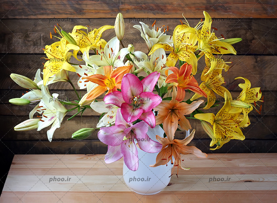 عکس با کیفیت گل های لیلیوم با رنگ های زیبا داخل گلدان سفید سفالی بر روی میز چوبی