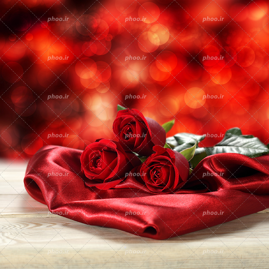 عکس با کیفیت سه شاخه گل رز قرمز بر روی پارچه ای قرمز بر روی میز چوبی و پس زمینه نورانی شده به رنگ قرمز