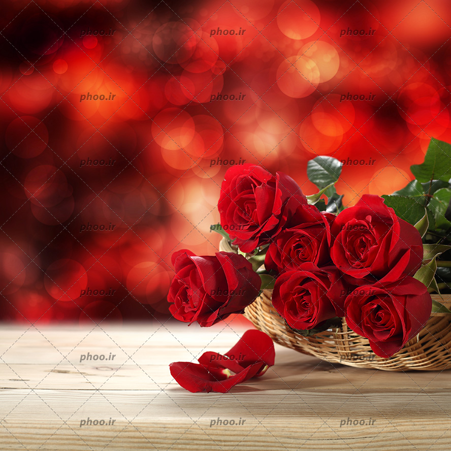 عکس با کیفیت دسته گل رز قرمز در سبد حصیری و چند گلبرگ بر روی میز چوبی و پس زمینه نورانیه رنگ قرمز