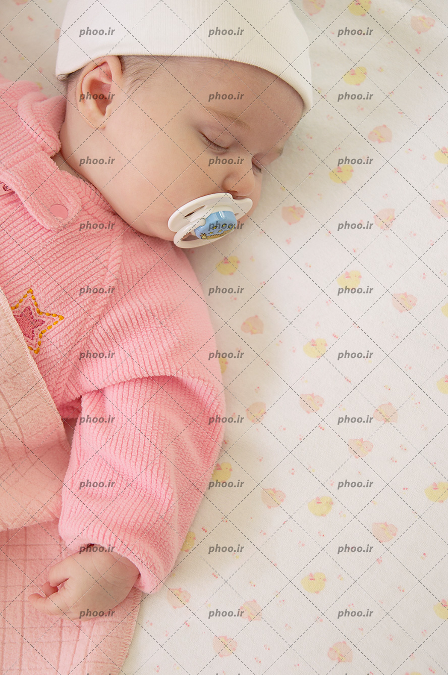 عکس با کیفیت نوزاد خواب با کلاه سفید و لباس صورتی و در حال پستونک خوردن