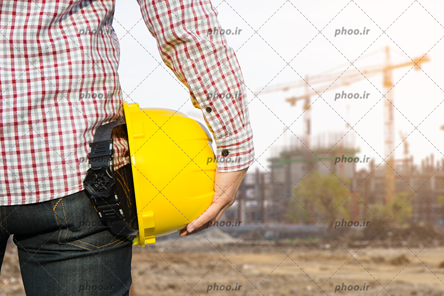 عکس با کیفیت مهندس عمران در حال نگاه کردن به ساختمان و کلاه ایمنی زرد رنگ او در دستانش
