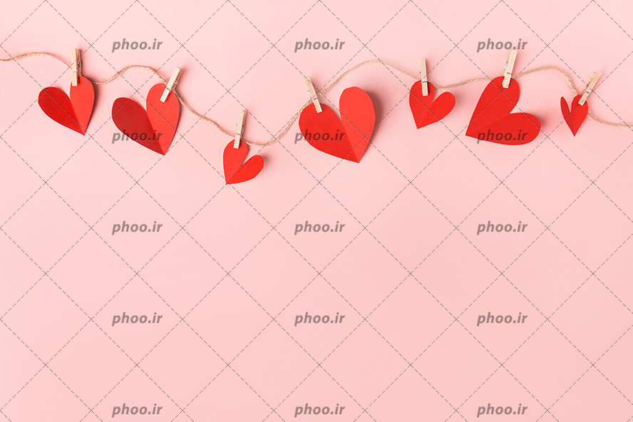 عکس با کیفیت قلب های قرمز کاغذی با گیره های چوبی کوچک وصل شده به نخ کنفی در پس زمینه صورتی