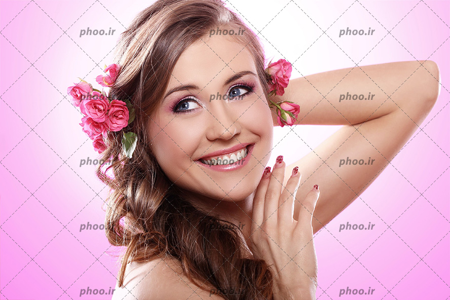 عکس با کیفیت زن با لبخند زیبا و موها به رنگ فندوقی و گل های رز صورتی در لا به لای موهایش و پس زمینه به رنگ صورتی
