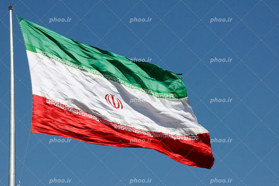 میله سفید و پرچم ایران وصل به آن در آسمان در حال تکان خوردن بر اثر وزش باد
