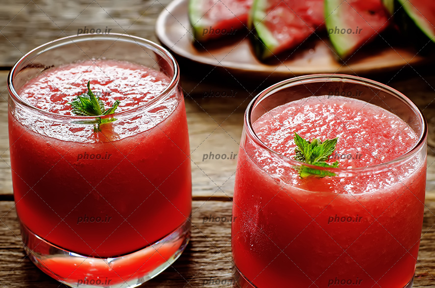 عکس با کیفیت دو لیوان آب هندوانه طبیعی و برگ های نعناع روی آنها بر روی میز چوبی و اسلایس های هندوانه در بشقاب چوبی در گوشه تصویر