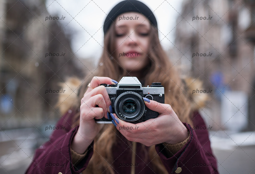عکس با کیفیت دختر عکاس زیبا با موهای بلوند و کلاهی مشکی بر سر و لباسی زرشکی در حال عکاسی در خیابان