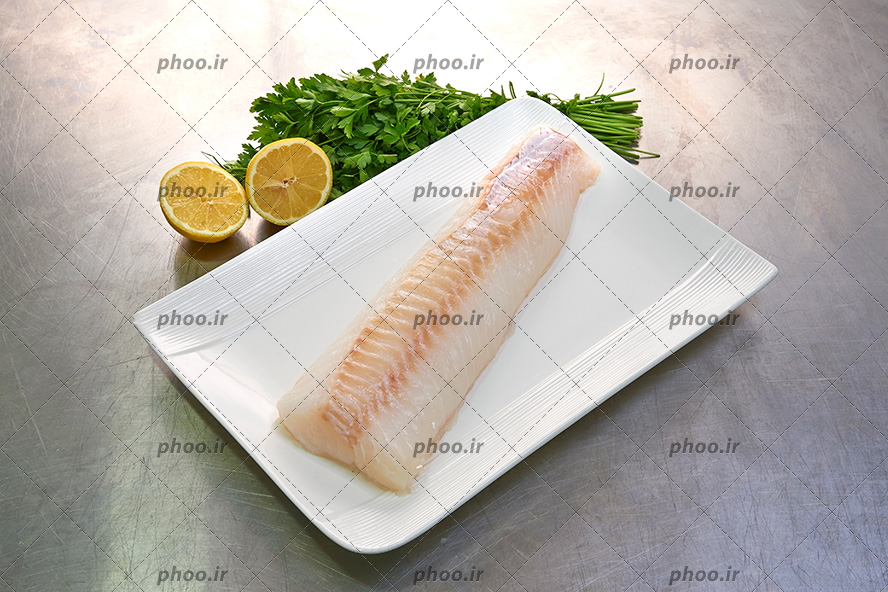 عکس با کیفیت قسمتی از ماهی پوست شده در ظرف سفید در کنار لیمو ترش ها و جعفری ها