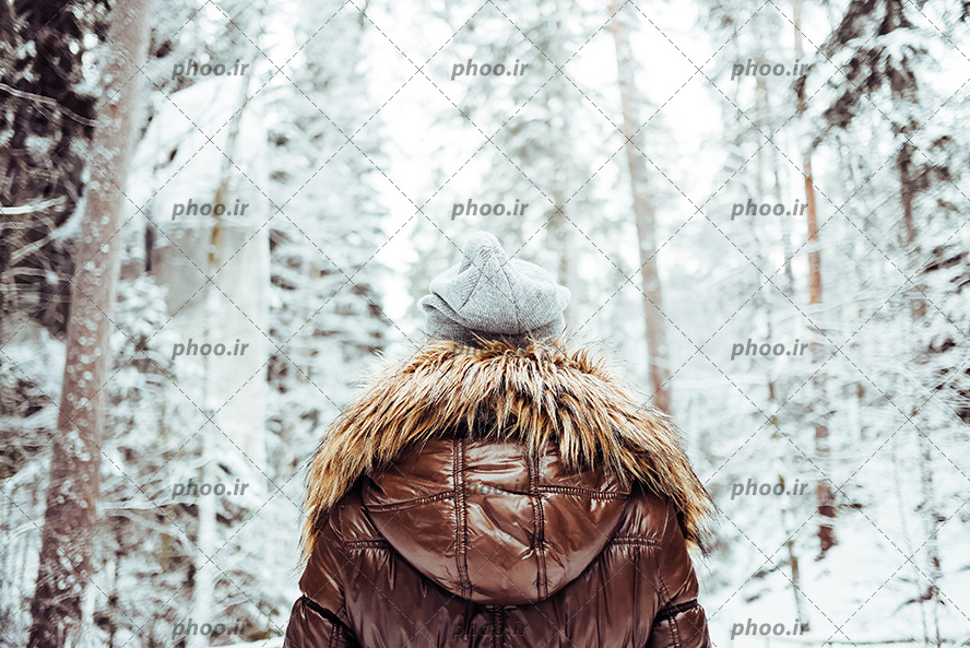 عکس با کیفیت دختر با کاپشن قهوه ای خز دار پشت به دوربین ایستاده و در جنگل برفی
