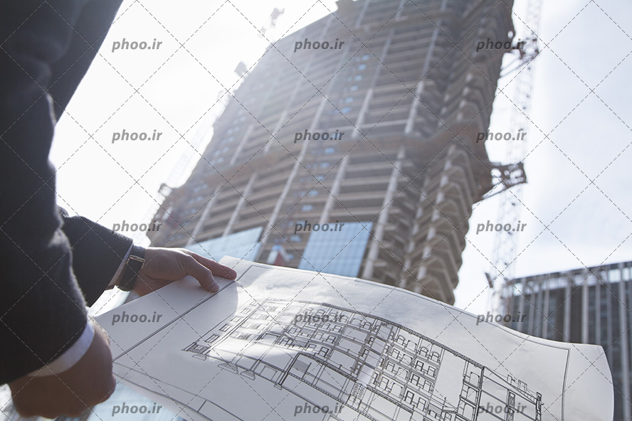 عکس با کیفیت مهندس عمران کت و شلوار پوش در حال نگاه کردن به نقشه در مقابل ساختمان نیمه ساز