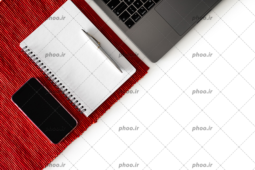 عکس با کیفیت دفتر فنری و روان نویس بر روی آن در کنار موبایل بر روی پارچه ی قرمز و لپ تاپ در گوشه تصویر