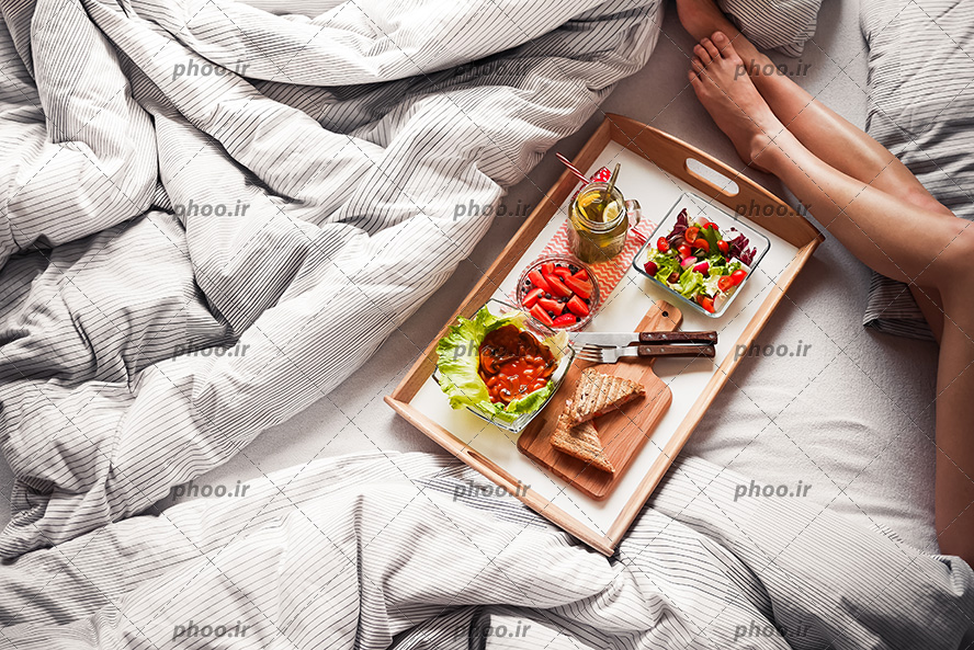 عکس با کیفیت سینی صبحانه در تخت خواب و پاهای زن در گوشه تصویر