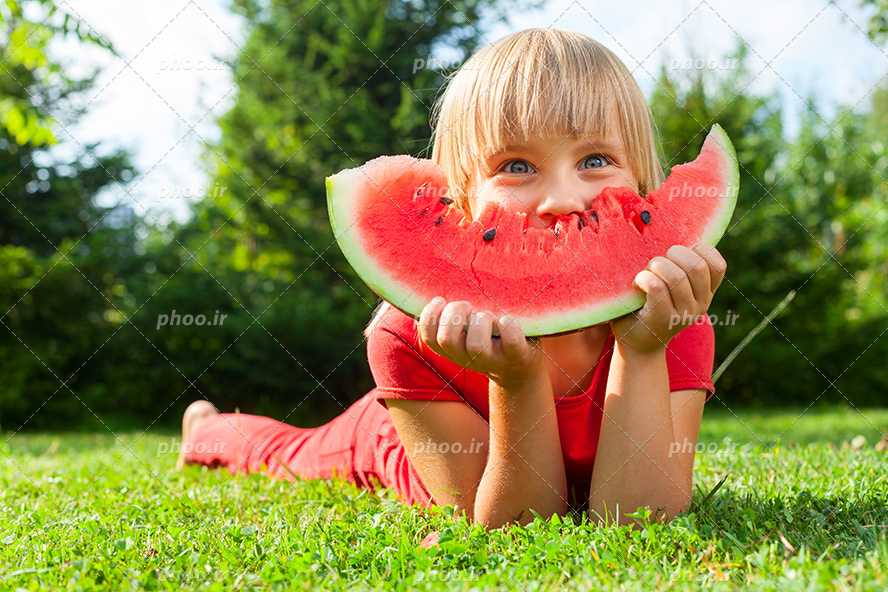عکس با کیفیت یک قاچ هندوانه ی قرمز در دست پسر بچه دراز کشیده در چمن های سرسبز