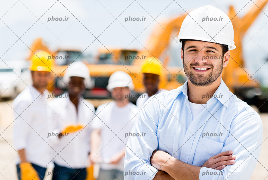 عکس با کیفیت مهندس با کلاه ایمنی و ایستاده با ژست و در حال لبخند زدن و چند مهندس ساختمان ایستاده در پشت سرش