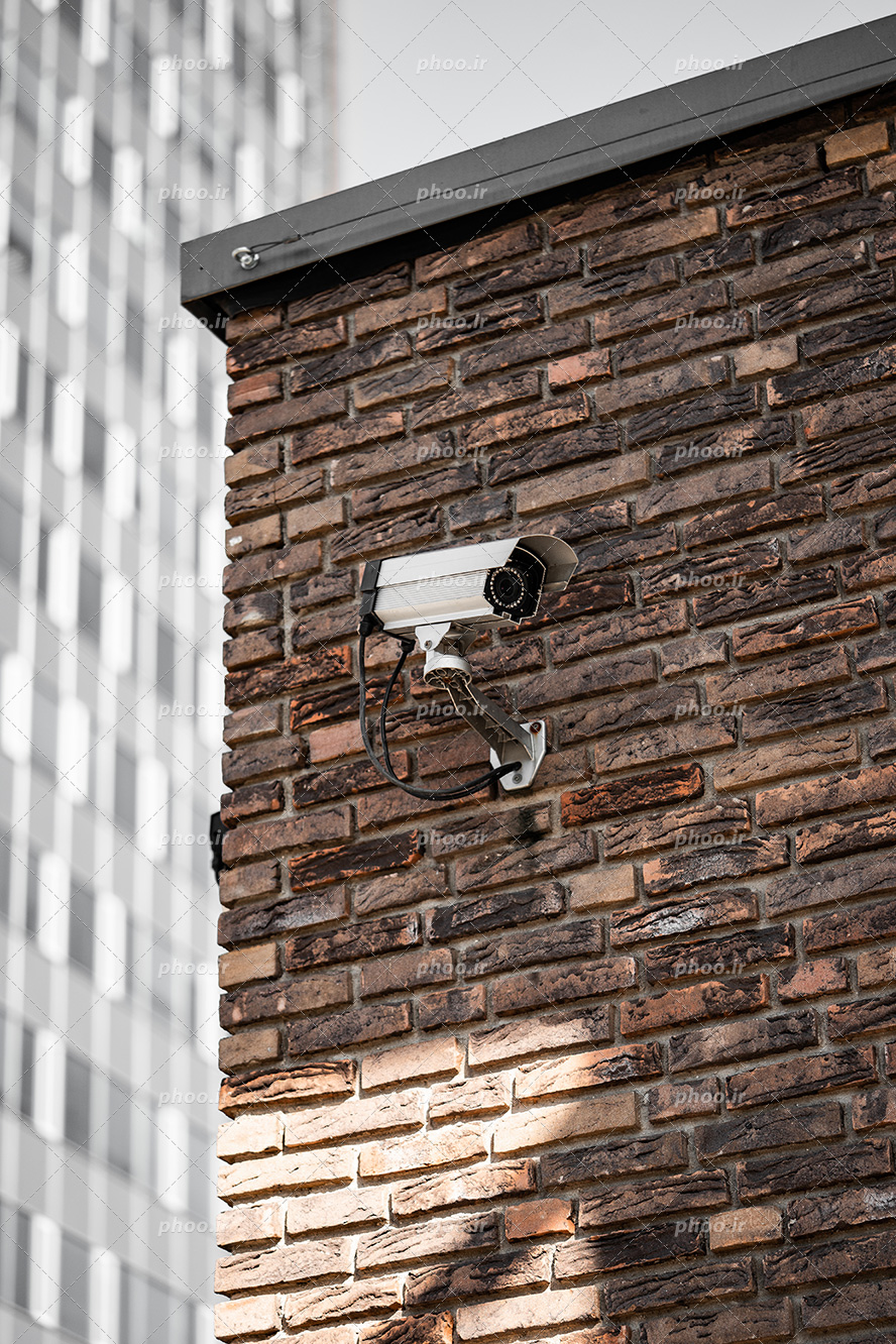 عکس با کیفیت دوربین مدار بسته در روی دیوار آجری و در گوشه تصویر ساختمان مرتفع