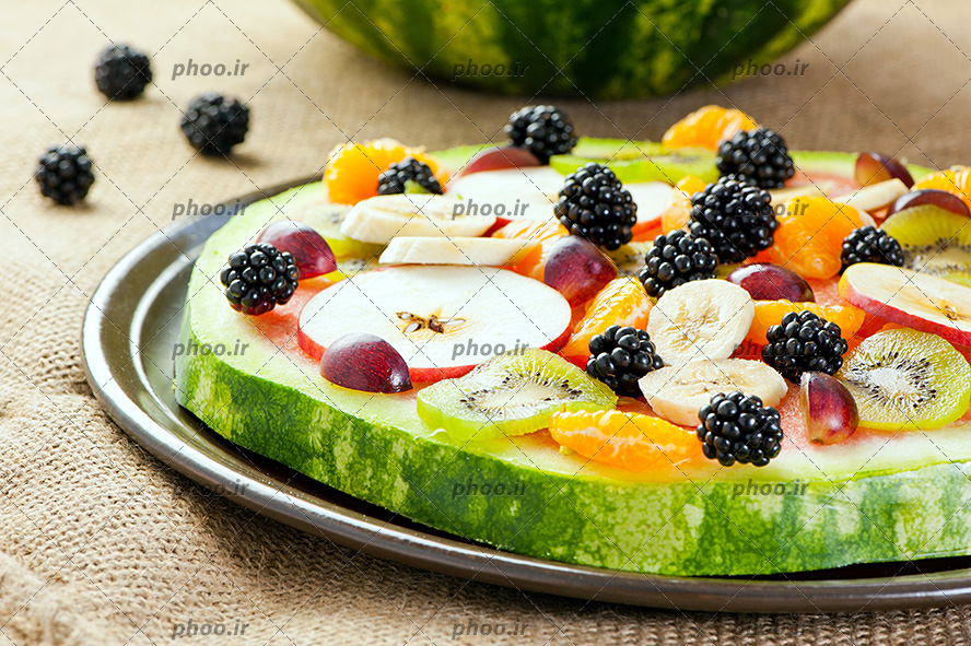 عکس با کیفیت اسلایس دایره ای هندوانه در سینی و میوه های نارنگی و کیوی و سیب و توت سیاه بر روی هندوانه