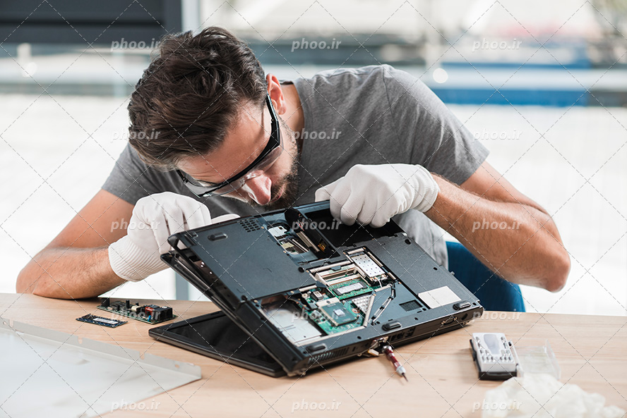 عکس با کیفیت مرد تعمیرکار با عینک و دستکش در حال باز کردن و تعمیر کردن لپ تاپ