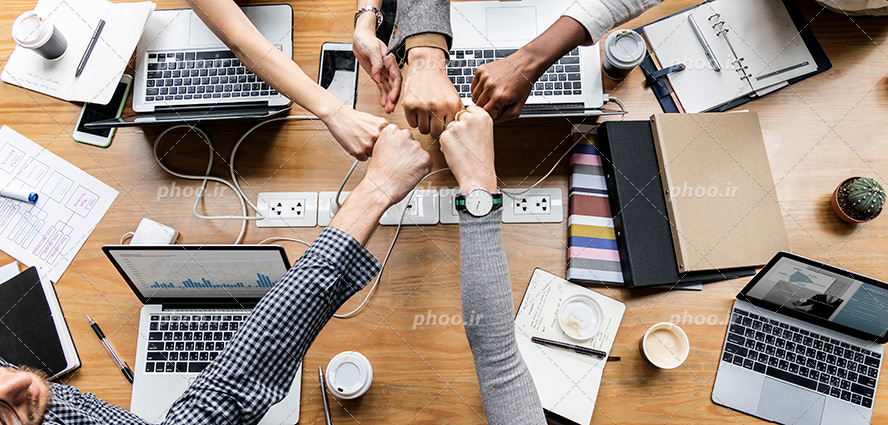 عکس با کیفیت شش کارمند نشسته در پشت میز و در حال مشت زدن به دست های یکدیگر به منظور پیروزی و موفقیت