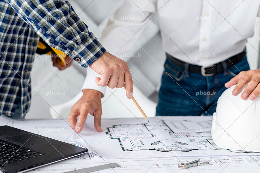 عکس با کیفیت مرد سفید پوش در حال توضیح دادن نقشه به مرد چارخونه پوش و لپ تاپ و کلاه ایمنی در تصویر