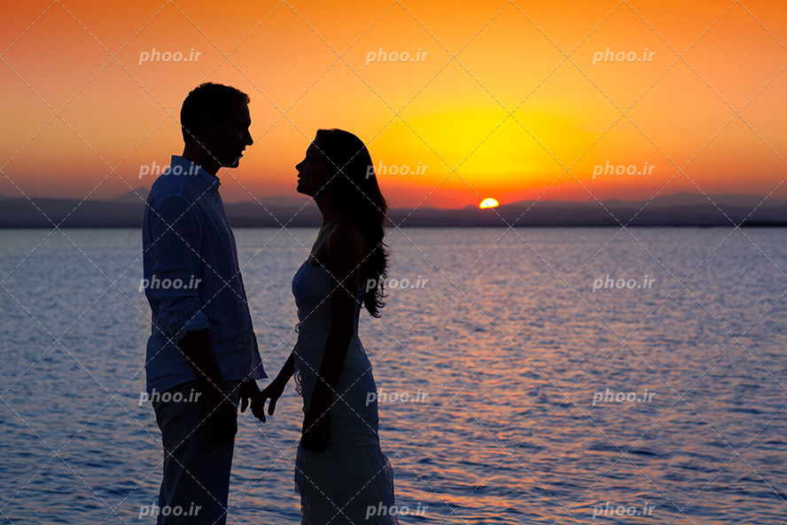 عکس با کیفیت زن و مرد در مقابل یکدیگر در کنار ساحل و خورشید در حال غروب و آسمان به رنگ نارنجی