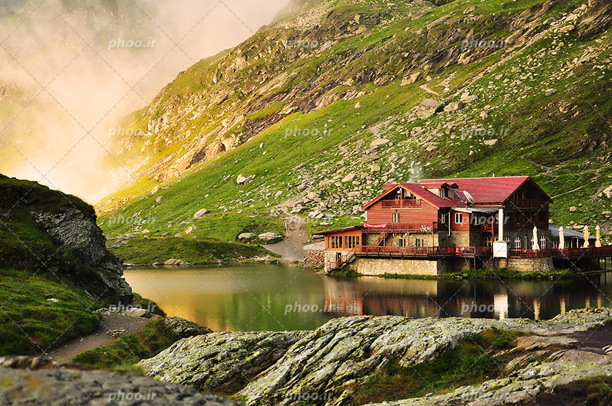 عکس با کیفیت خانه ی زیبا با سقف شیروانی به رنگ قرمز در دامنه ی کوه ها و در کنار دریاچه کوچک و طبیعت زیبای سرسبز