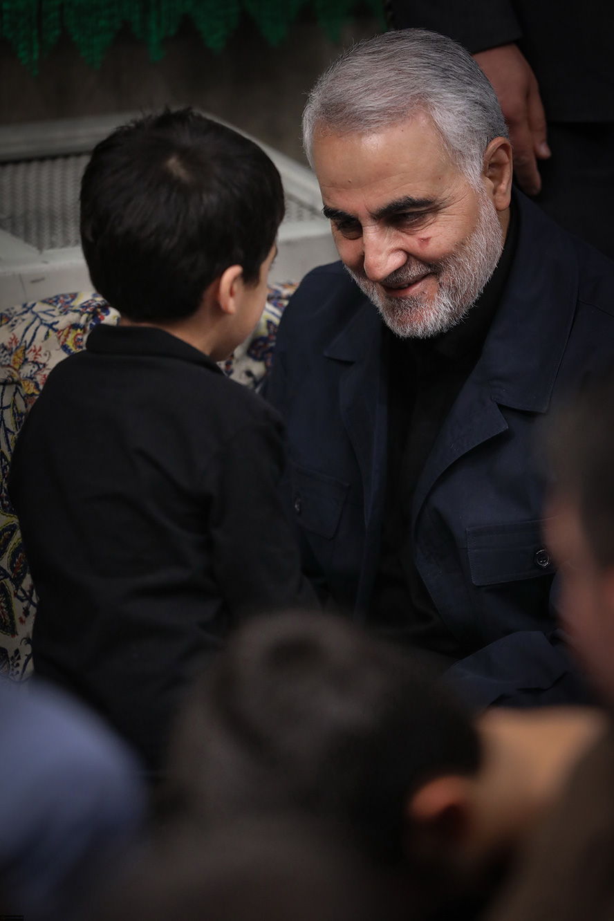 عکس با کیفیت تصویر شهید سردار قاسم سلیمانی در حال صحبت کردن با خنده با پسر بچه کوچک با لباس مشکی بر تنش