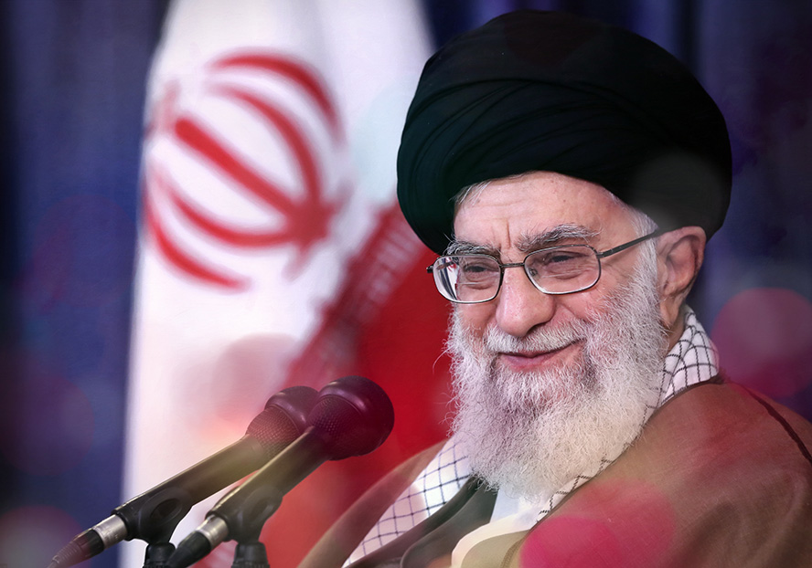 عکس با کیفیت حضرت خامنه ای در حال لبخند زدن با عبا قهوه ای و عمامه مشکی و پرچم ایران در کنار ایشان