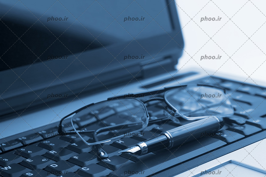 عکس لپ تاپ خاموش و عینک و خودنویس روی صفحه کلید لپ تاپ