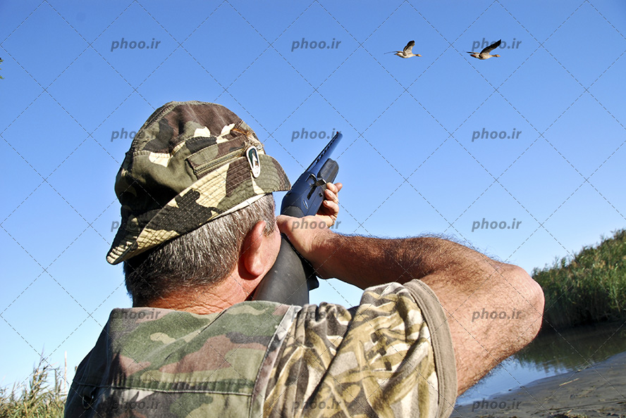 عکس با کیفیت مرد شکارچی با اسلحه در حال شکار پرنده یا مرغابی در کنار برکه یا دریاچه با لباس پلنگی