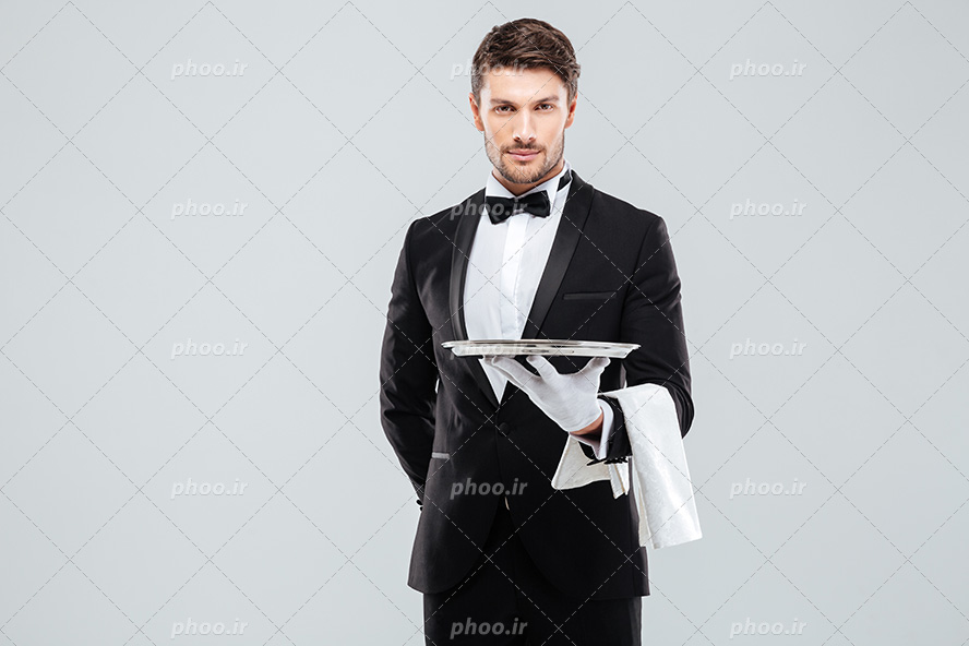 عکس با کیفیت گارسون با کت و شلوار و پاپیون مشکی و سینی در دست و پارچه سفید بر روی ساعد دستش