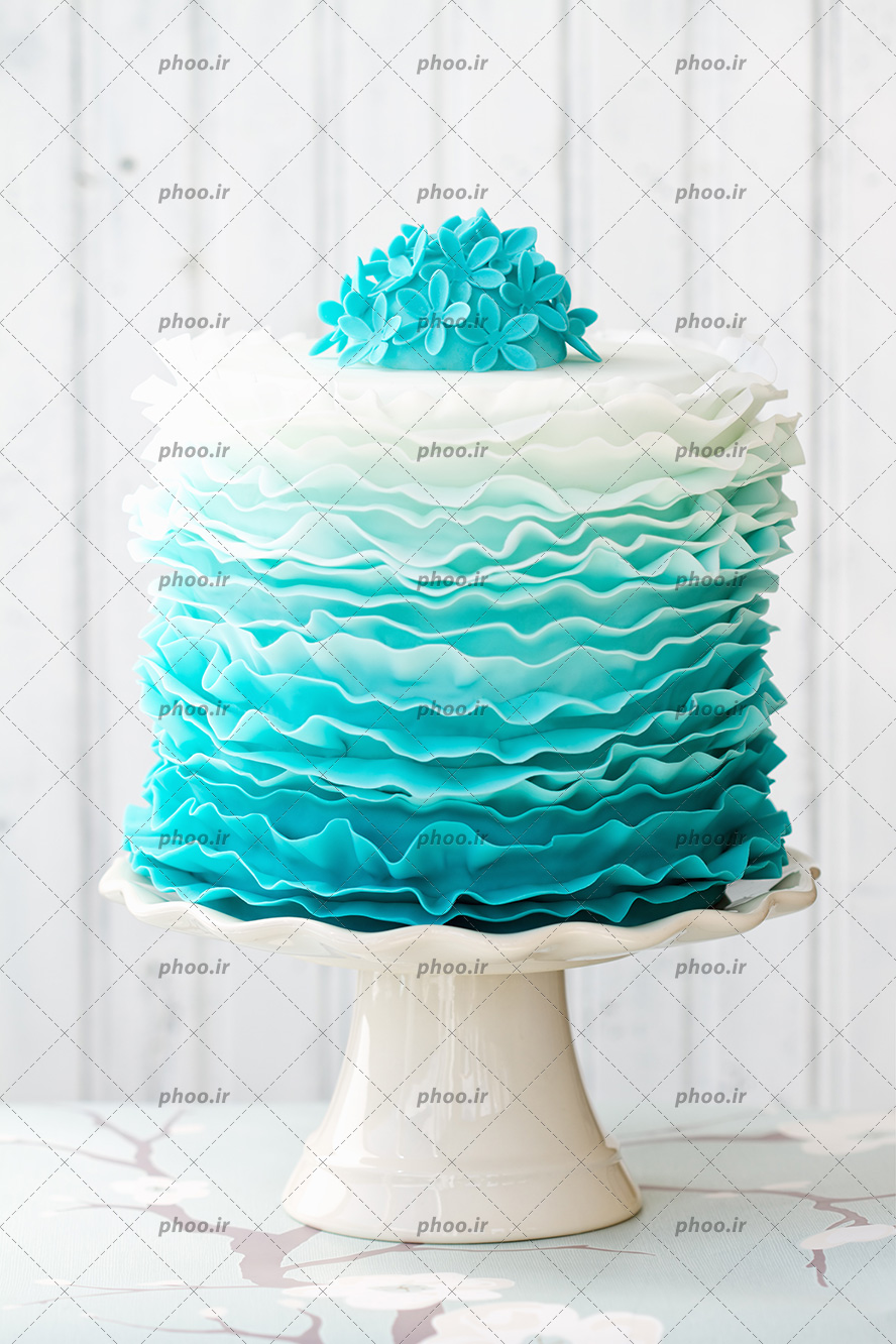 عکس با کیفیت کیک زیبا به رنگ آبی فیروزه ای که کم کم رنگ آن به سفید نزدیک شده است و تزئین شده با گل های فیروزه ای کوچک در بالا ی کیک و در ظرف مخصوص قرار گرفته است