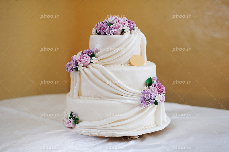 عکس با کیفیت کیک سه طبقه سفید تزئین شده با گل های بنفش زیبا و برگ های سبز