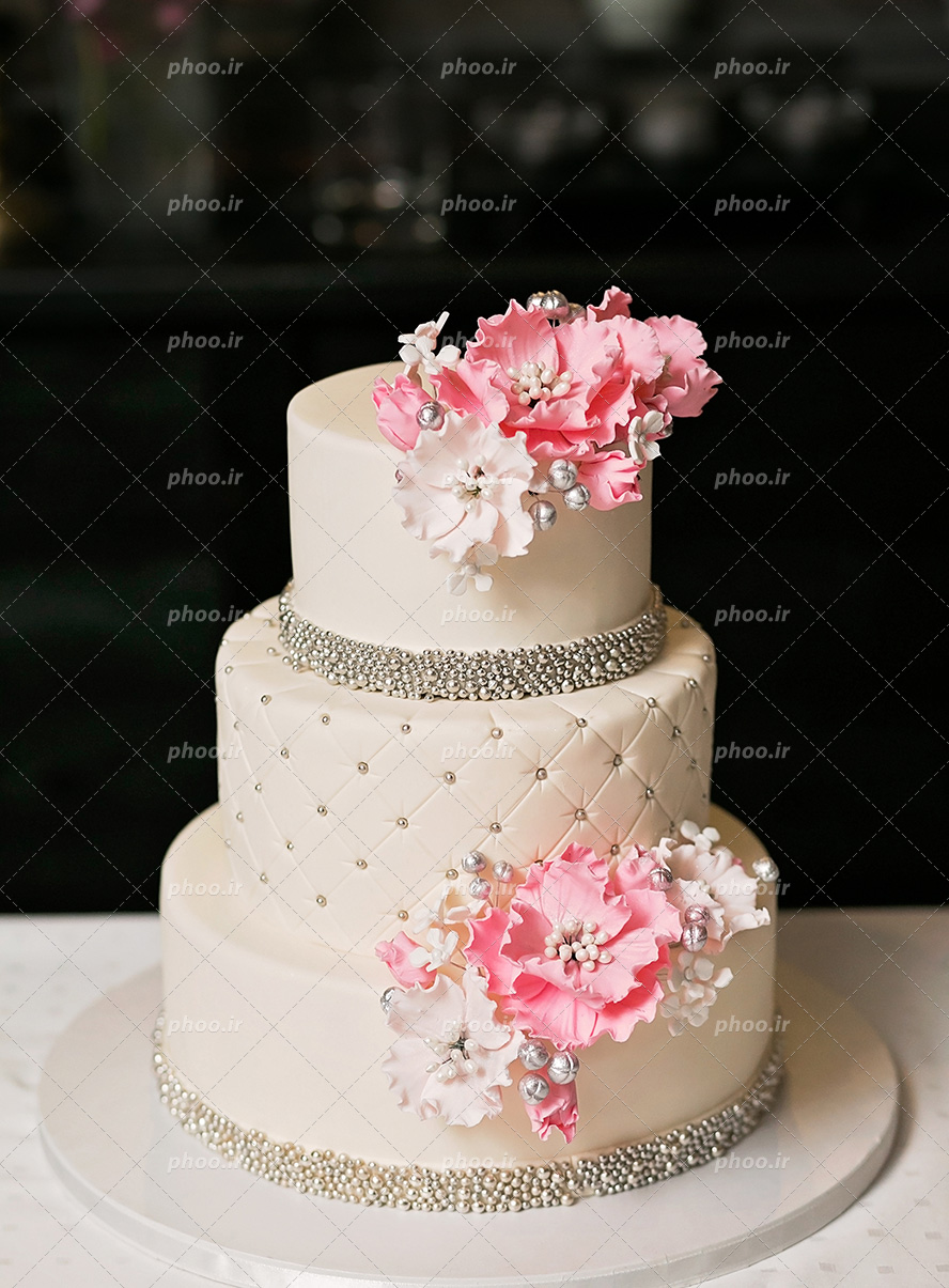 عکس با کیفیت کیک سه طبقه سفید تزئین شده با گل های صورتی و مروارید های خوراکی نقره ای