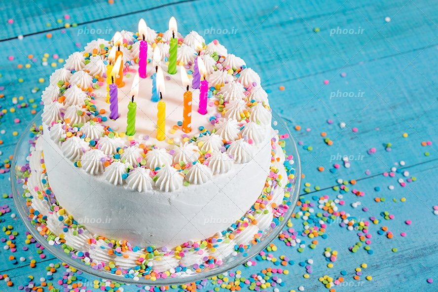 عکس با کیفیت کیک خامه ای تزئین شده با ترافل های رنگی و شمع های رنگارنگ و قرار گرفته بر روی میز چوبی آبی