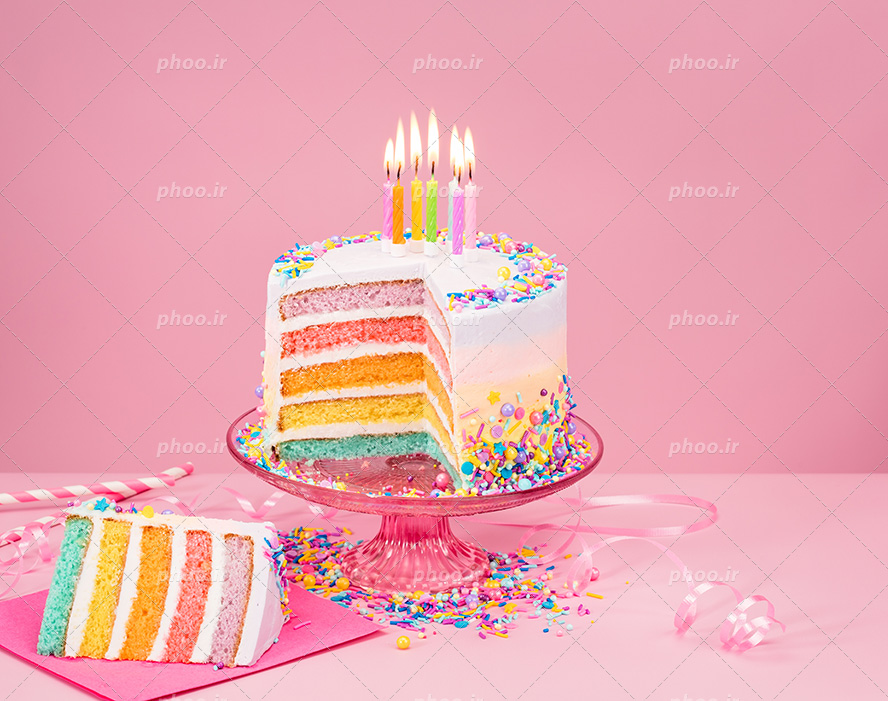 عکس با کیفیت کیک خامه ای تزئین شده با ترافل های رنگی و شمع های رنگارنگ و داخل کیک به شکل رنگین کمان و قرار گرفته بر روی استند کیک در بک گراند صورتی