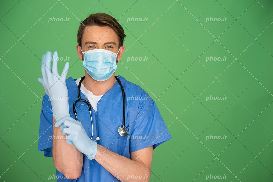 عکس با کیفیت پزشک با روپوش و ماسک آبی در حال دست کردن دستکش آبی و پس زمینه به رنگ سبز