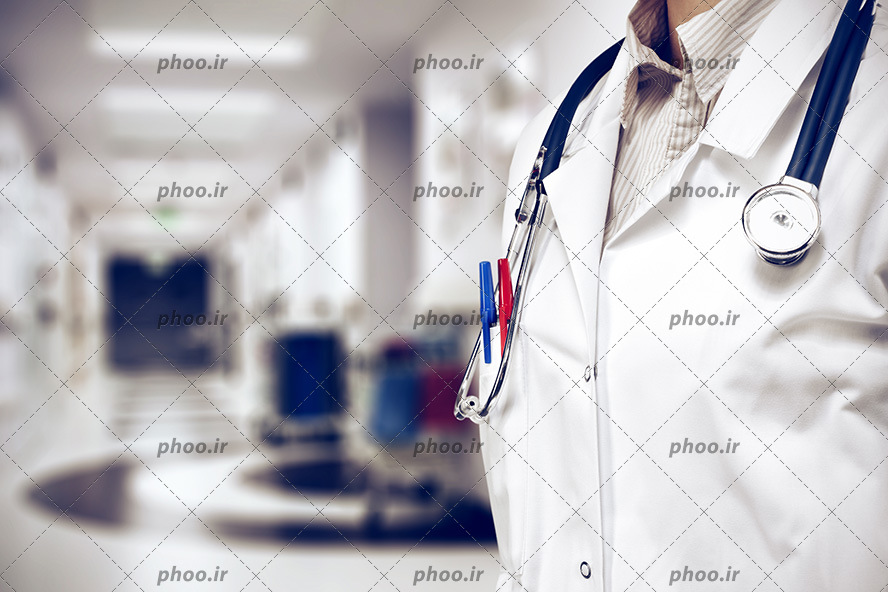 عکس با کیفیت پزشک ایستاده در راه روی بیمارستان و روپوش سفید به تن و استتوسکوپ بر روی گردن و دو خودکار در جیب