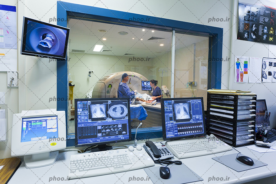 عکس با کیفیت کامپیوتر ها و پرونده های بیماران بر روی میز و پنجره شیشه ای بر روی دیوار اتاق و آن طرف پنجره اتاق سیتی اسکن و دو پزشک در کنار دستگاه سیتی اسکن