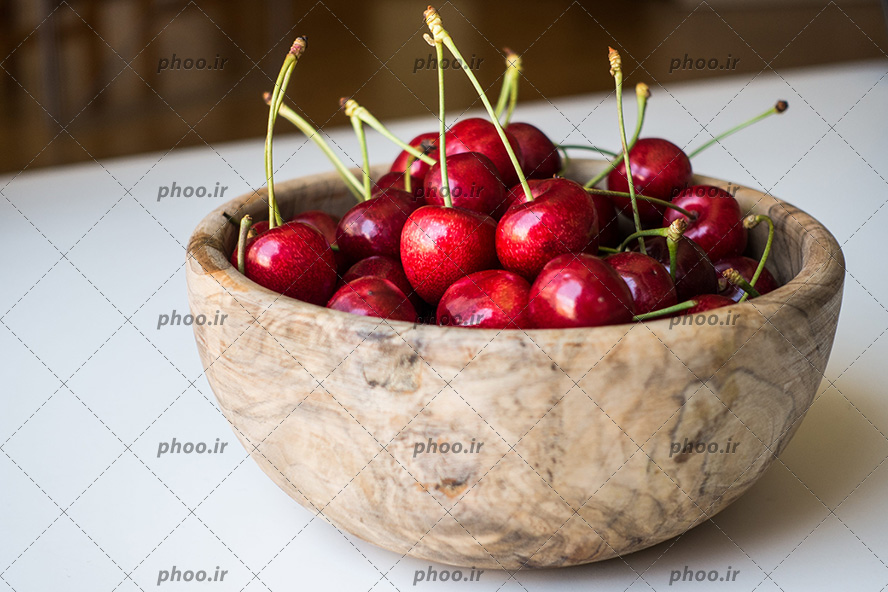 عکس با کیفیت گیلاس های قرمز و خوش رنگ و قرمز داخل ظرف چوبی بر روی میز سفید