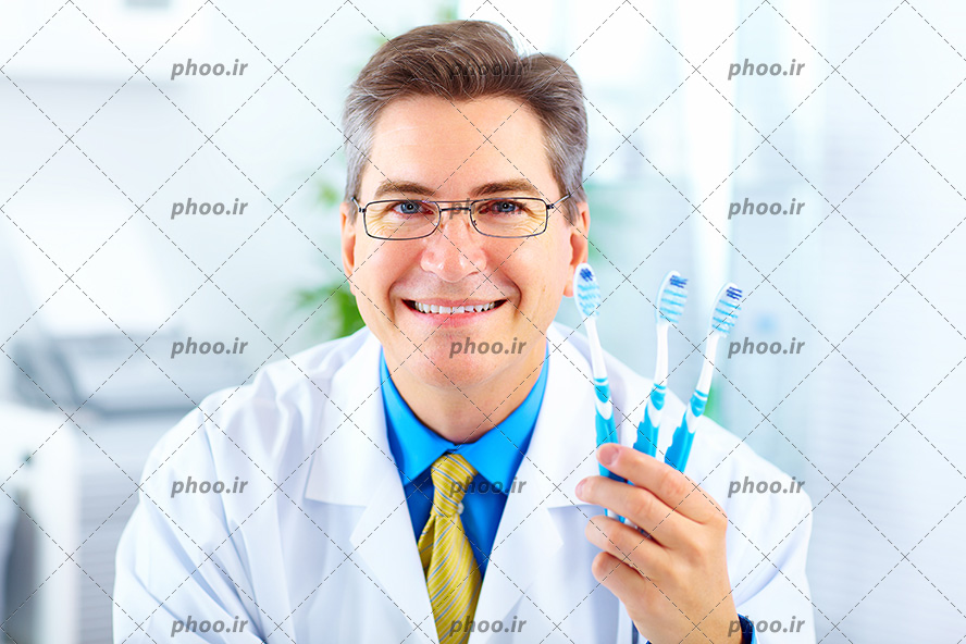 عکس با کیفیت سه مسواک در دست پزشک و پزشک لبخند بر لب و با روپوش سفید و پیراهن آبی و کروات زرد