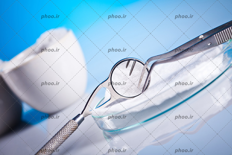عکس با کیفیت پنس و آینه مقعر دندانپزشکی بر روی ظرف شیشه ای پر شده از دسمال های استریل و بک گراند به رنگ آبی